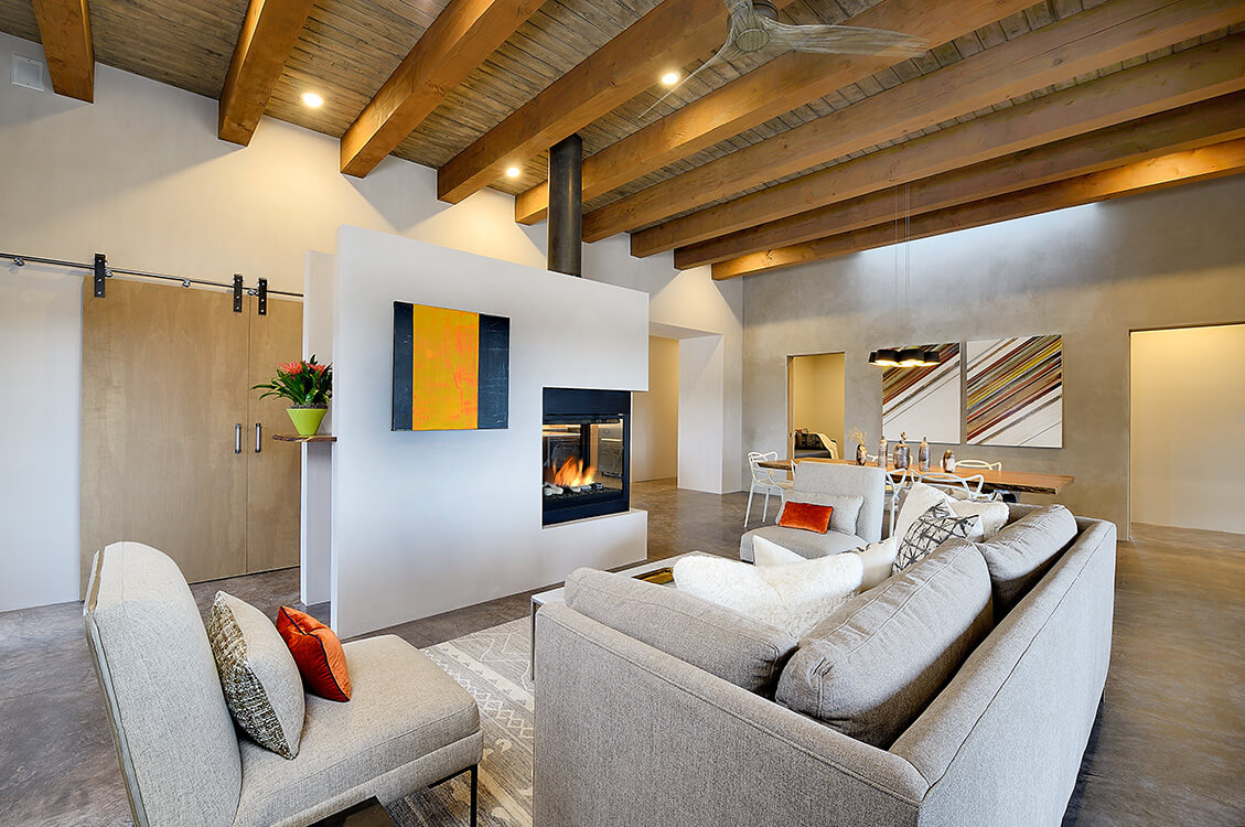 Modern living room with exposed wooden ceiling beams, a central double-sided fireplace, neutral-toned sofas, contemporary art, and a dining area in the background.