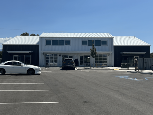 A modern commercial building with large windows and a metal roof. Three cars are parked in front. One person stands nearby. The parking area has a handicap accessible space. Blue sky overhead.