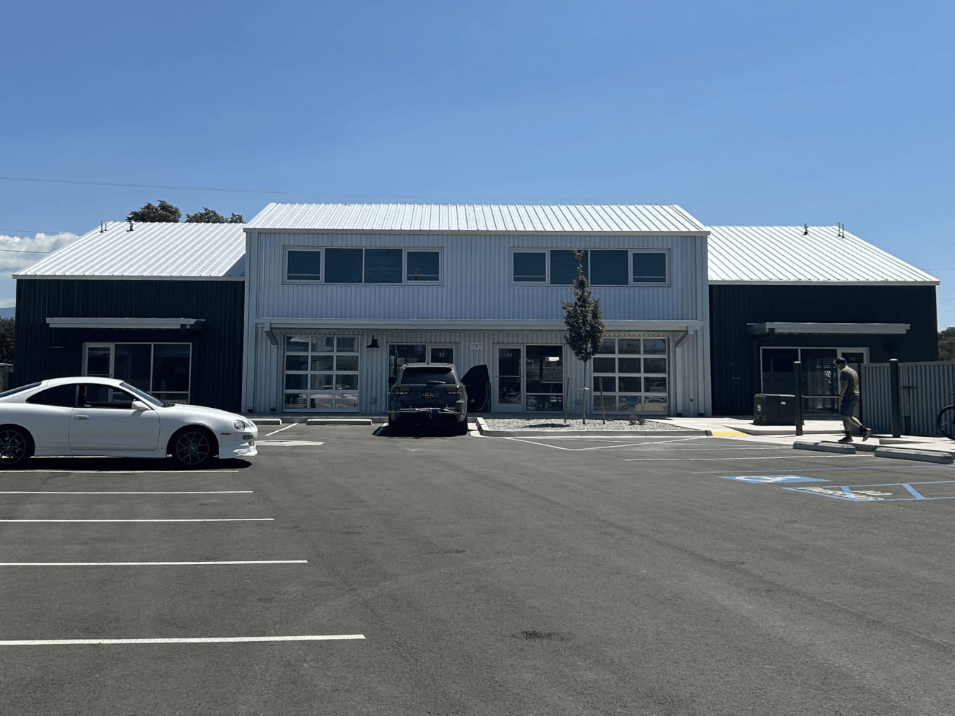 A modern commercial building with large windows and a metal roof. Three cars are parked in front. One person stands nearby. The parking area has a handicap accessible space. Blue sky overhead.