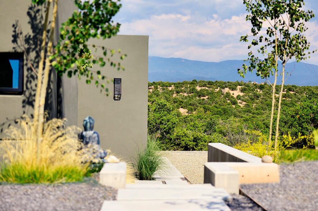 Modern house entrance with concrete pathway, desert landscaping, a small Buddha statue, and a view of green hills and distant mountains under a partly cloudy sky.