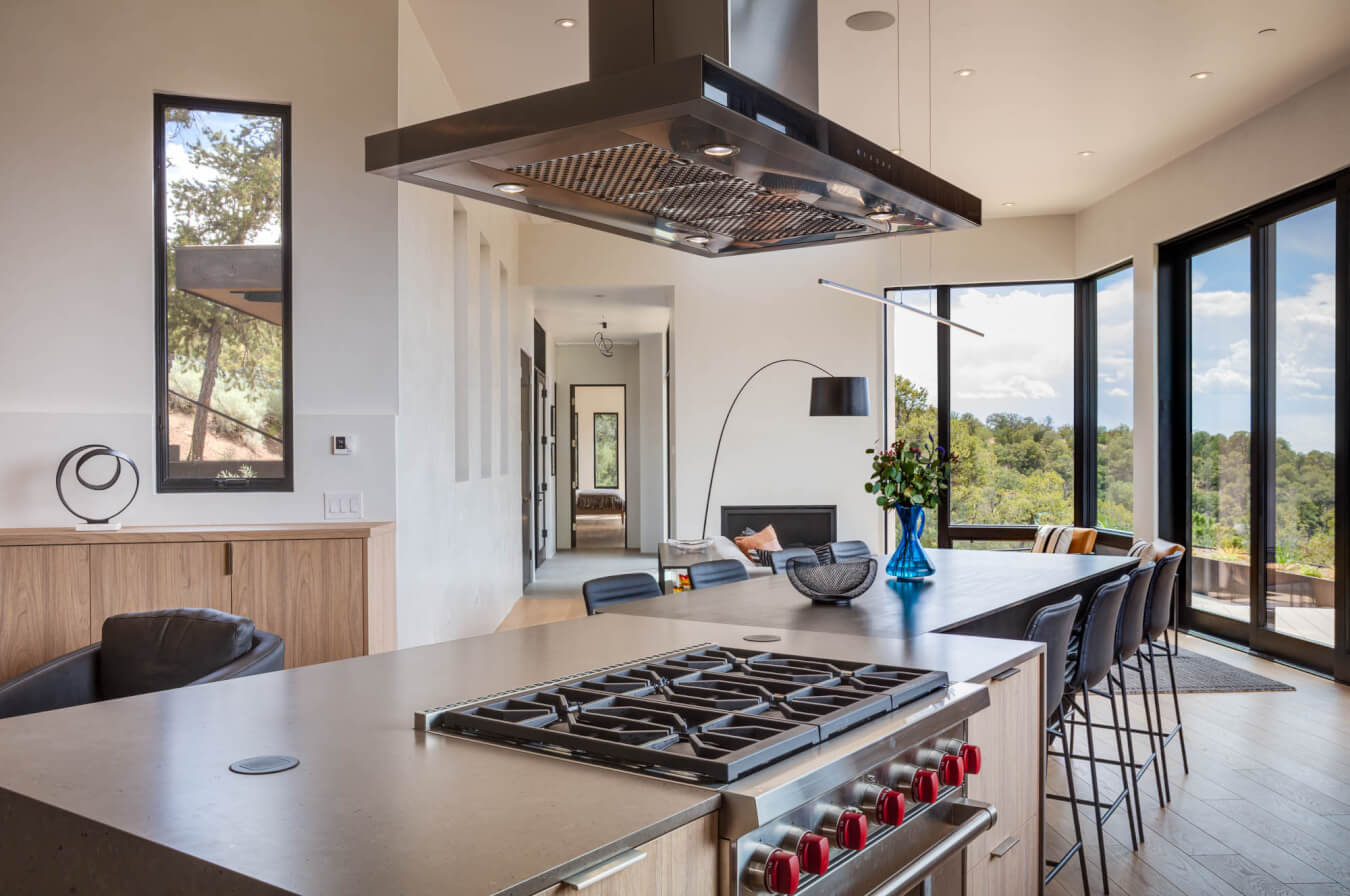 Modern kitchen with a large island, gas stove, bar stools, living area, and floor-to-ceiling windows overlooking a scenic outdoor view.