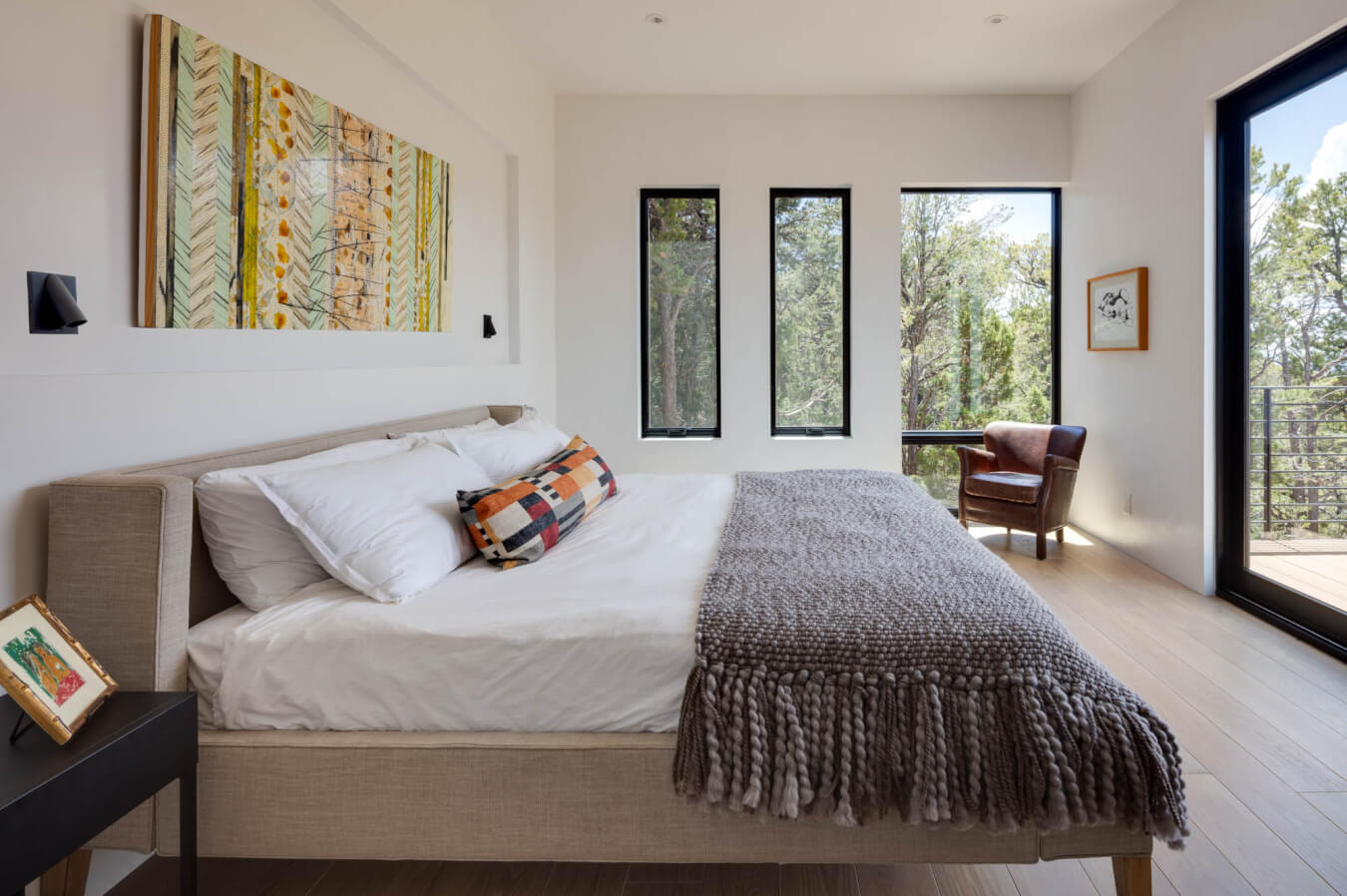 A modern bedroom with a neatly made bed, textured gray blanket, patterned pillow, artwork above the headboard, large windows, an armchair, and a view of trees outside.