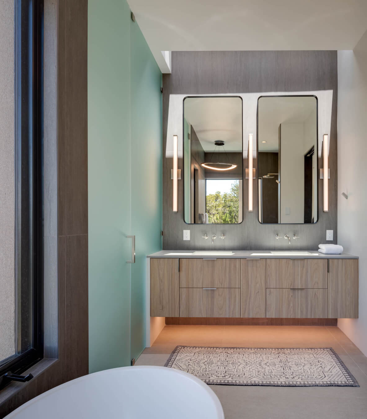 Modern bathroom with double sinks, two rectangular mirrors, vertical wall lights, floating wood vanity, frosted glass door, and a window providing natural light.