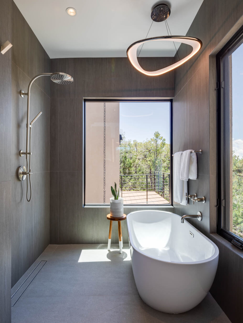 Modern bathroom with a freestanding bathtub, large window, rainfall shower, towel rack, and a small plant on a wooden stool.