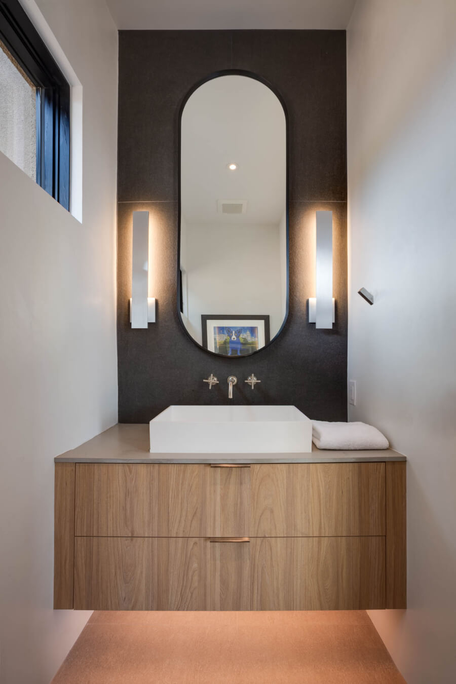 Modern bathroom with a floating wood vanity, rectangular vessel sink, oval mirror, two wall sconces, towel, and artwork reflected in the mirror.