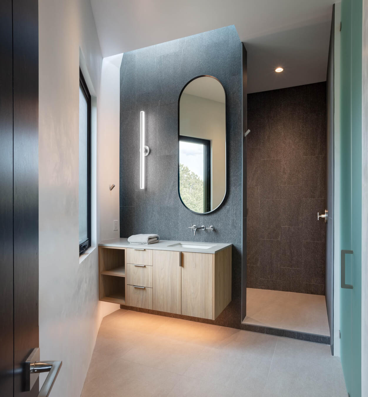 Modern bathroom with a floating wood vanity, oval mirror, vertical light fixture, large window, and a walk-in shower with dark tile walls.