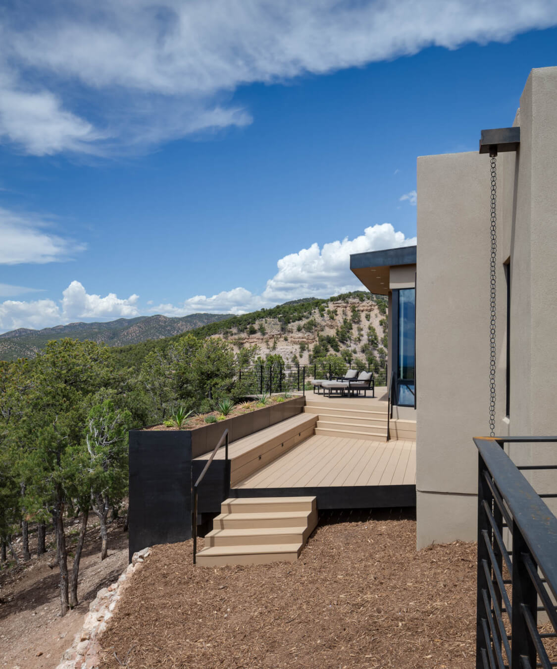 Modern house with a beige exterior and a wooden deck overlooking a forested hillside under a blue sky with scattered clouds.