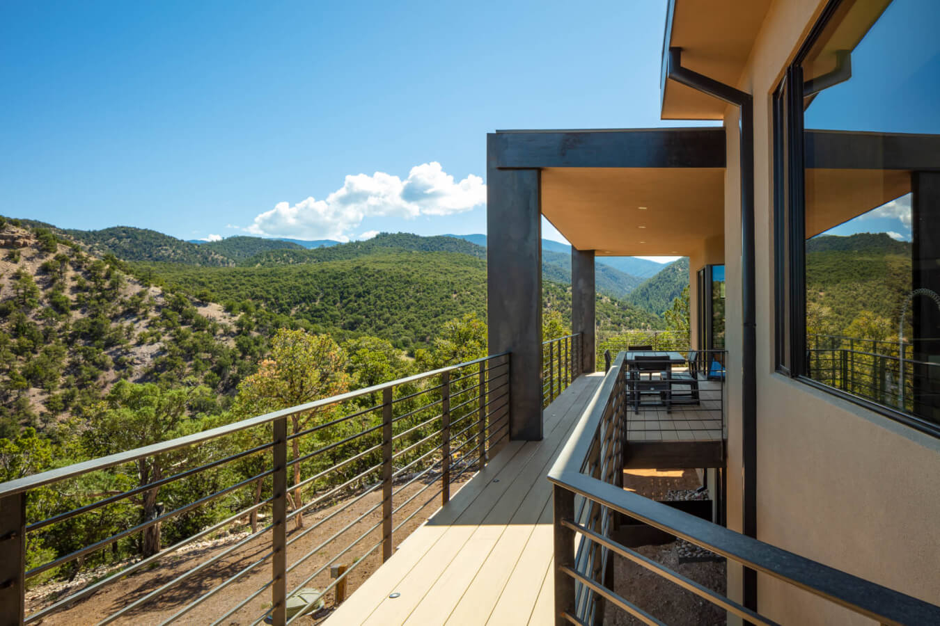 A modern house balcony with metal railings overlooking a green, mountainous landscape under a clear blue sky.