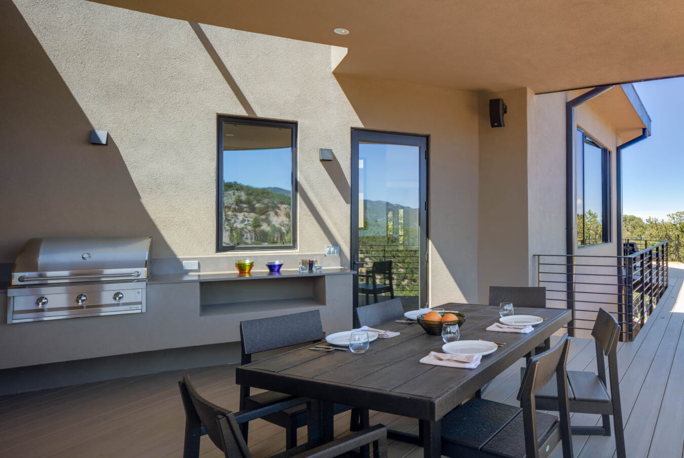 Modern outdoor patio with a dining table set for four, a built-in stainless steel grill, and views of trees and hills through glass windows and doors.