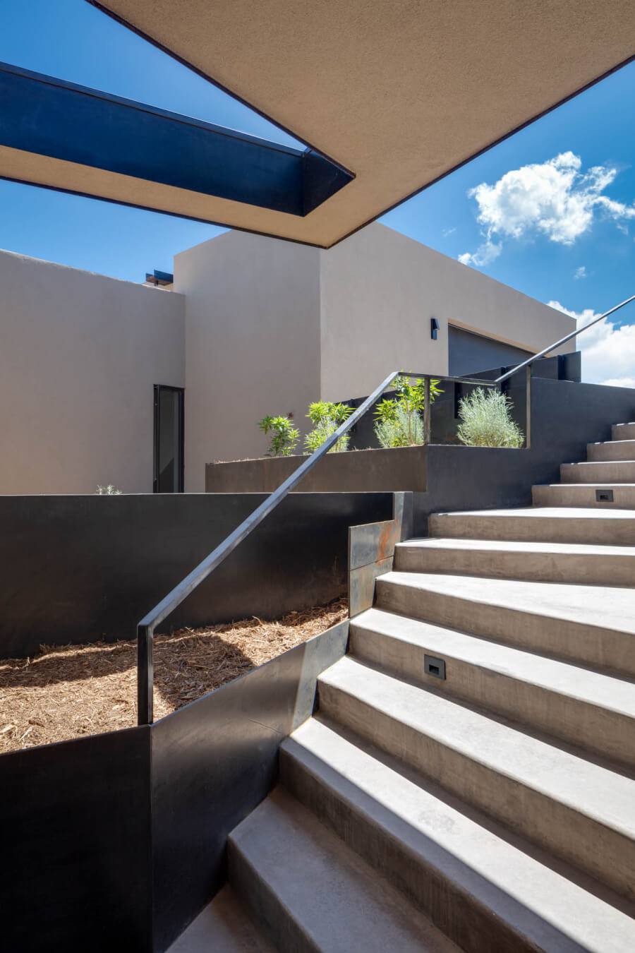 Concrete stairs with a metal handrail lead to a modern building with beige walls, minimalist planters, and a skylight above. Blue sky with clouds in the background.