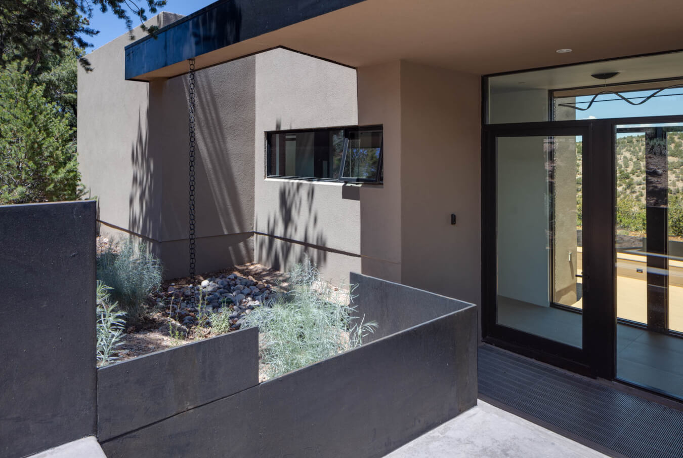 Modern house entrance with a concrete planter, rain chain, glass door, and surrounding natural vegetation.