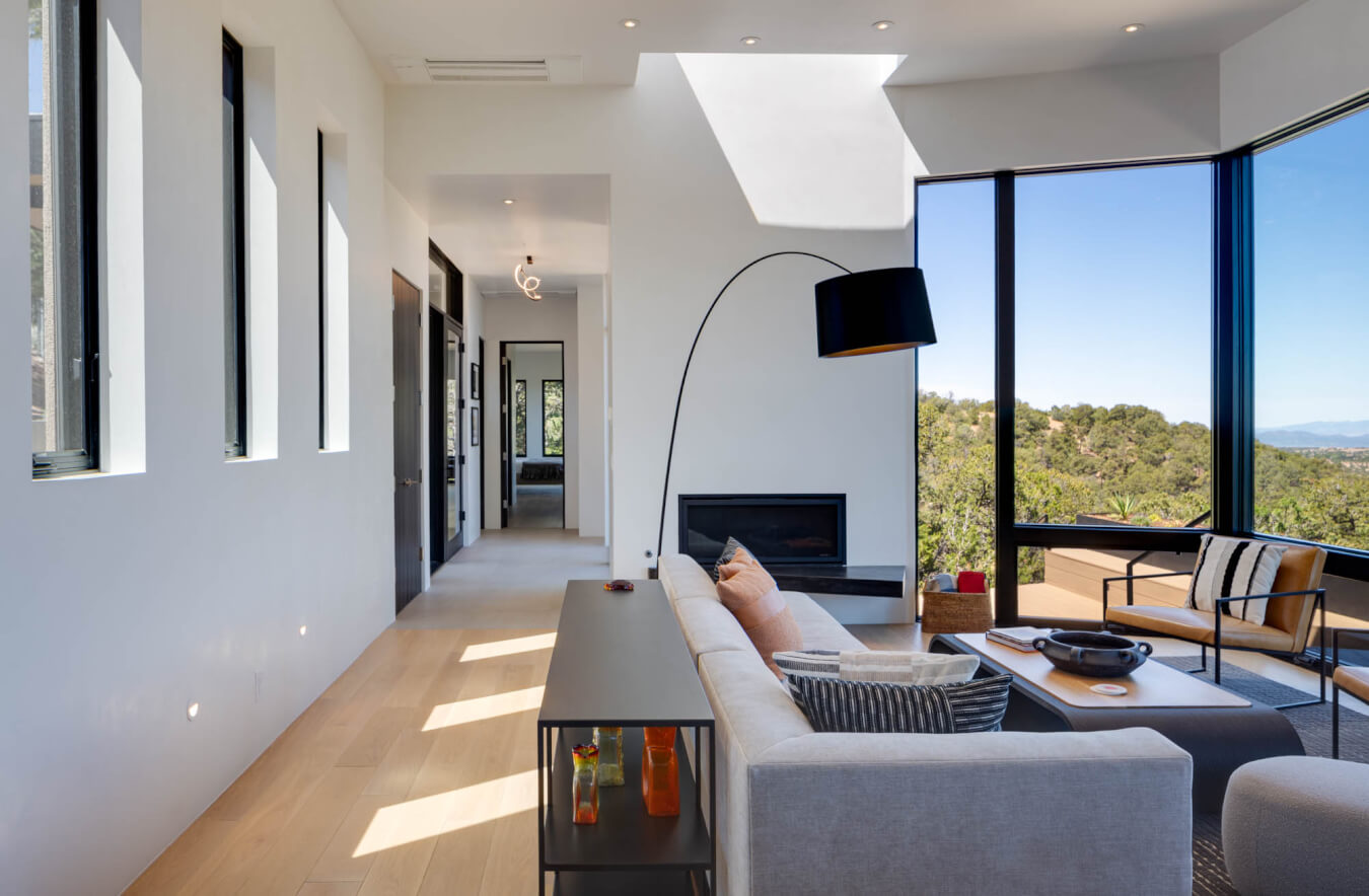 Modern living room with a gray sectional sofa, black floor lamp, fireplace, and large windows overlooking outdoor scenery; a hallway leads to other rooms.