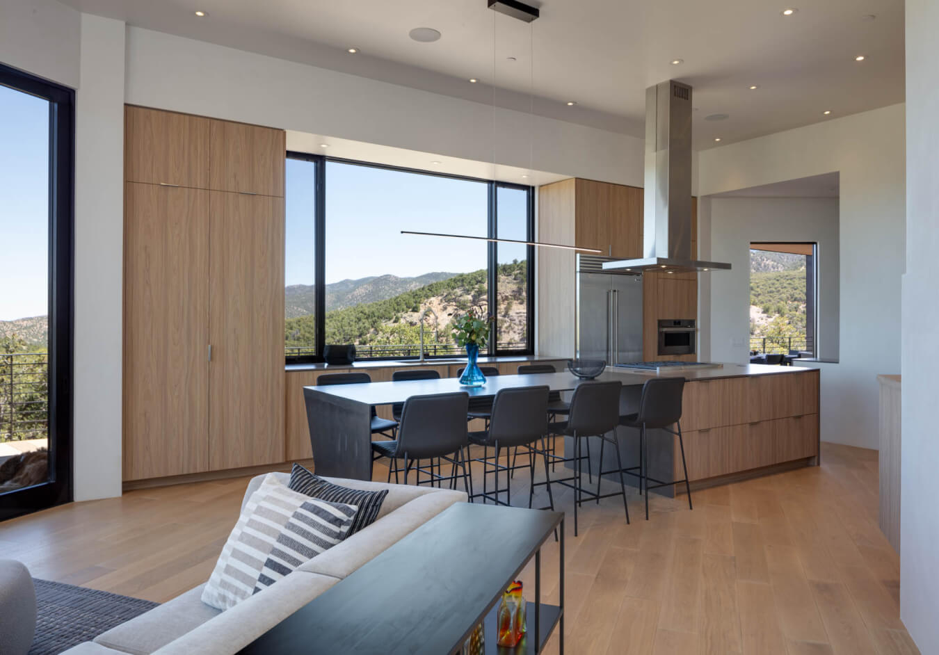 Modern open-plan kitchen and dining area with light wood cabinets, large windows showing mountain views, a long table with black chairs, and a gray sofa in the foreground.