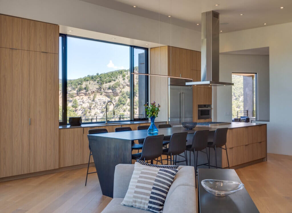 Modern kitchen with wood cabinets, a large island with black seating, built-in appliances, and expansive windows offering a view of hills and trees.
