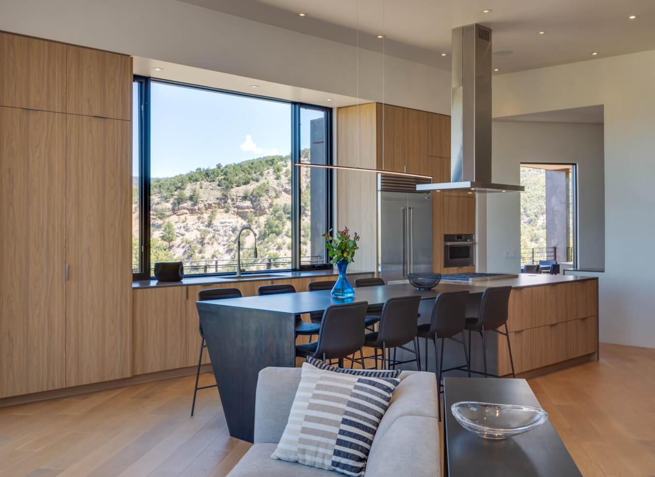 Modern kitchen with wood cabinets, a large island with black seating, built-in appliances, and expansive windows offering a view of hills and trees.