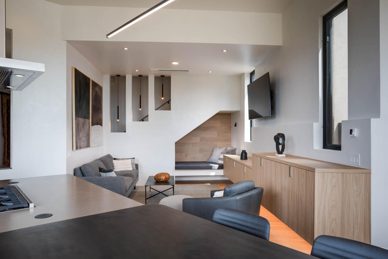 Modern open-plan living room with minimalist decor, light wood cabinetry, gray sofas, built-in bench nook, wall-mounted TV, and abstract art on the wall.