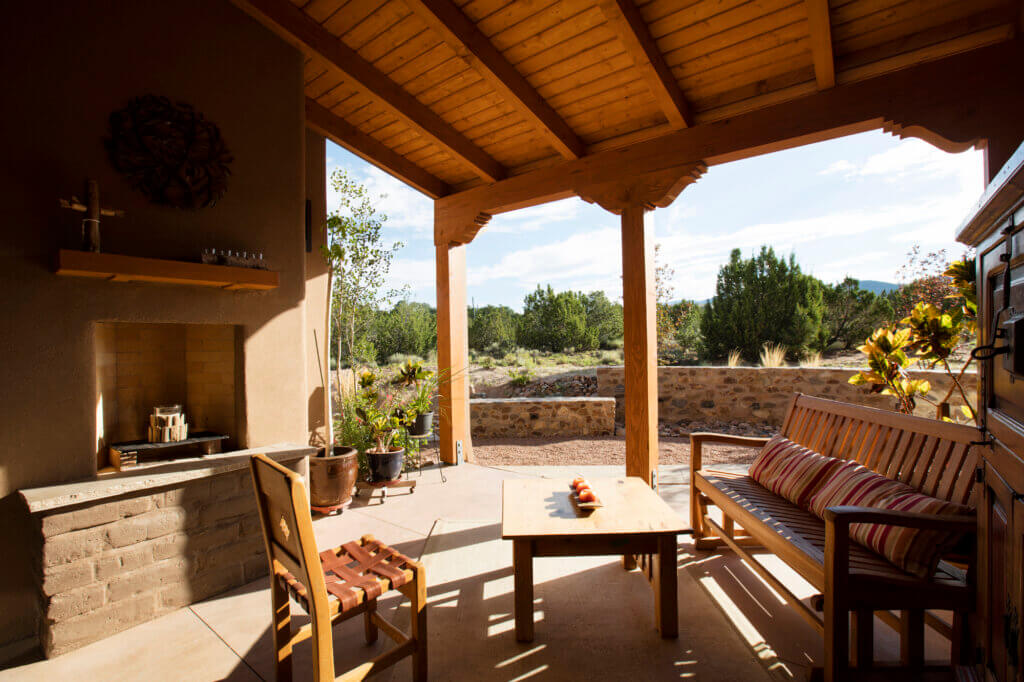 Covered patio with wooden furniture, a fireplace, and potted plants, overlooking a garden with trees and a stone wall under a sunny sky.