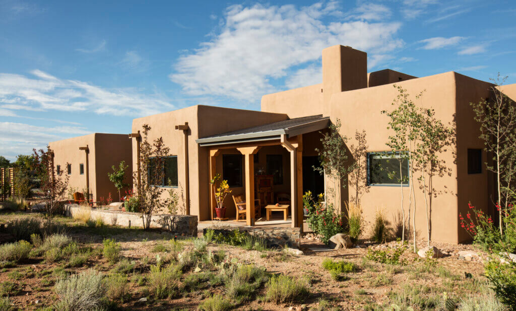 A modern adobe-style house with flat roofs, tan stucco walls, and a covered front porch, surrounded by dry landscaping and desert plants under a blue sky.
